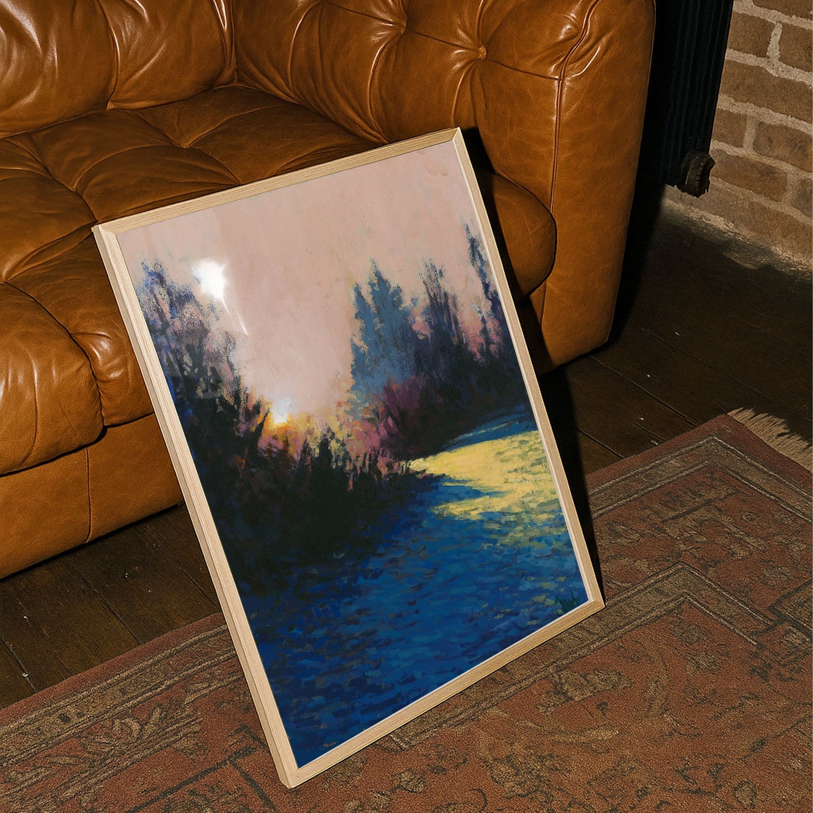 Painting of a sunset over water on a wooden easel against a brown leather couch and brick wall.